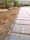 A close-up view of a paved pathway with rectangular stone tiles partially covered in dirt. The path leads towards a driveway with parked cars visible in the background. Green plants and bushes line the path on one side.