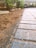 A close-up view of a paved pathway with rectangular stone tiles partially covered in dirt. The path leads towards a driveway with parked cars visible in the background. Green plants and bushes line the path on one side.
