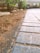 A close-up view of a paved pathway with rectangular stone tiles partially covered in dirt. The path leads towards a driveway with parked cars visible in the background. Green plants and bushes line the path on one side.