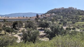 A panoramic view of a historic site with ancient ruins on a hill, surrounded by lush greenery and trees. In the background, there is a large structure that appears to be a classic example of ancient architecture. The landscape includes well-maintained pathways and open spaces, set against a backdrop of distant mountains and a clear blue sky.