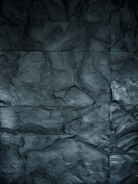 A textured surface featuring large, irregular stone tiles with varying shades of dark gray. The surface has an uneven, rocky appearance with visible grooves and contours that mimic natural stone.