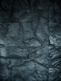 A textured surface featuring large, irregular stone tiles with varying shades of dark gray. The surface has an uneven, rocky appearance with visible grooves and contours that mimic natural stone.