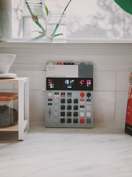 A compact electronic music sampler sits on a white kitchen countertop. Two glass jars with cut stems are placed on the windowsill above. A coffee container with a wooden lid is on a small shelf to the left, alongside part of a white ceramic bowl. The background features an out-of-focus view through a window, suggesting a serene, indoor scene.