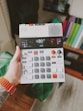 A hand holds a compact electronic music device featuring various buttons, dials, and a small screen. The person wearing an orange wristband has a tattoo on their arm. The background includes a blurred indoor setting with books or folders visible on a shelf.