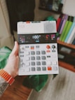 A hand holds a compact electronic music device featuring various buttons, dials, and a small screen. The person wearing an orange wristband has a tattoo on their arm. The background includes a blurred indoor setting with books or folders visible on a shelf.