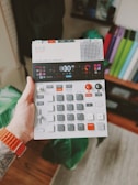 A hand holds a compact electronic music device featuring various buttons, dials, and a small screen. The person wearing an orange wristband has a tattoo on their arm. The background includes a blurred indoor setting with books or folders visible on a shelf.