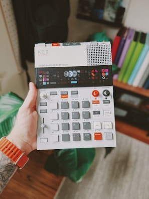 A hand holds a compact electronic music device featuring various buttons, dials, and a small screen. The person wearing an orange wristband has a tattoo on their arm. The background includes a blurred indoor setting with books or folders visible on a shelf.