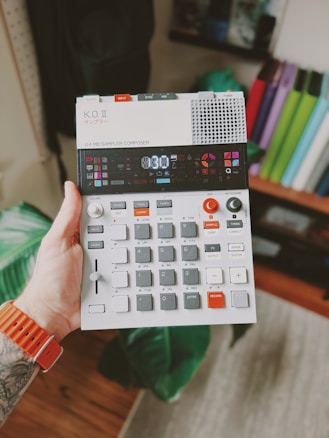 A hand holds a compact electronic music device featuring various buttons, dials, and a small screen. The person wearing an orange wristband has a tattoo on their arm. The background includes a blurred indoor setting with books or folders visible on a shelf.