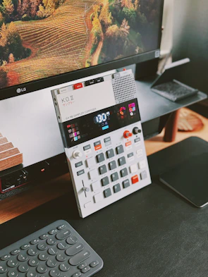 A modern electronic music sampler and composer device with various buttons and a digital display is placed on a desk. The background features a large computer monitor displaying an image of a lush, terraced vineyard. A sleek wireless keyboard and a black mousepad are also visible.