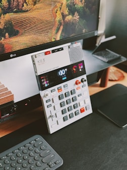 A modern electronic music sampler and composer device with various buttons and a digital display is placed on a desk. The background features a large computer monitor displaying an image of a lush, terraced vineyard. A sleek wireless keyboard and a black mousepad are also visible.