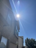 A bright sun emits strong rays over a residential building with a brick exterior. The sun creates a lens flare across the clear blue sky. The building features large windows and a distinct architectural design. A tree with full foliage is visible near the house, casting a shadow.