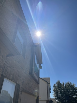 A bright sun emits strong rays over a residential building with a brick exterior. The sun creates a lens flare across the clear blue sky. The building features large windows and a distinct architectural design. A tree with full foliage is visible near the house, casting a shadow.