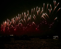 A night sky filled with bright, colorful fireworks being launched upwards and creating sparkling trails. The lower part of the image shows silhouettes of structures, possibly buildings or ships, against a dark backdrop.