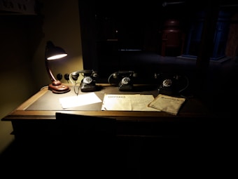 A vintage wooden desk is dimly lit by a classic table lamp with a curved neck. There are three old-fashioned rotary dial telephones aligned on the desk. Several papers and documents are spread out under the lamp&rsquo;s light, casting shadows across the surface. The surrounding area is dark and shadowy, suggesting an atmosphere of secrecy or historical significance.