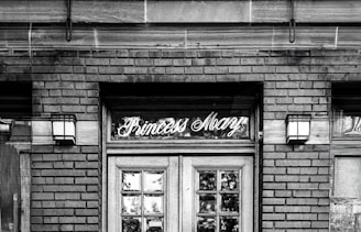 Elegant vintage signage above a wooden door on a brick building with two wall-mounted lanterns on either side. The name 'Princess Mary' is written in cursive script in the glass section above the door.