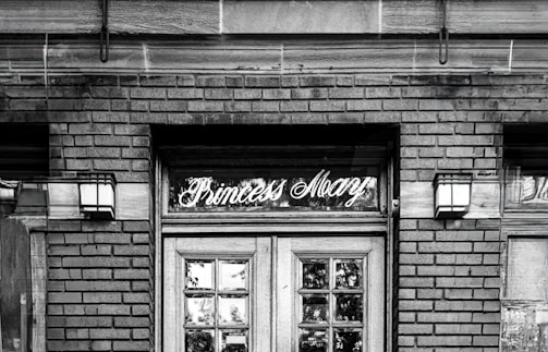 Elegant vintage signage above a wooden door on a brick building with two wall-mounted lanterns on either side. The name 'Princess Mary' is written in cursive script in the glass section above the door.