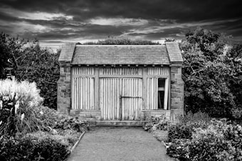 A small, weathered wooden shed with stone accents stands surrounded by dense foliage. The sky is overcast, creating a moody atmosphere. The shed's doors have vertical slats, and the structure appears aged, hinting at a rustic, rural setting. Lush plants and grasses flank the pathway leading to the shed.