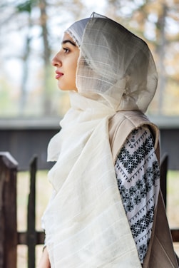 A person is wearing a light, semi-transparent headscarf with intricate embroidery on their clothing. The background is softly blurred, emphasizing the subject.