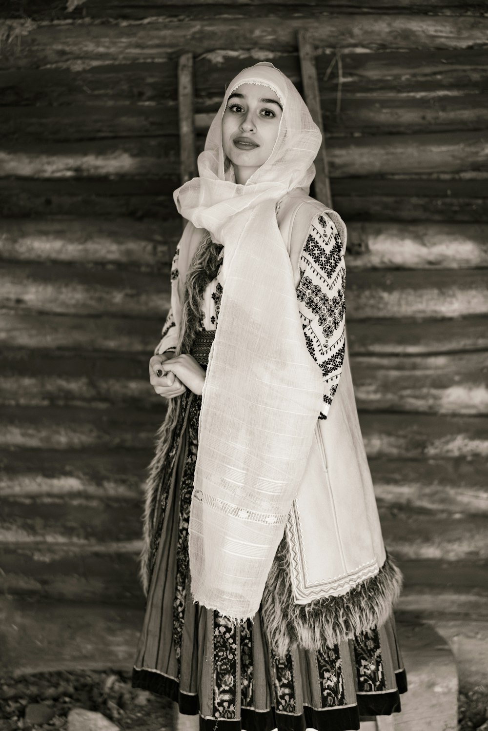 a woman in a dress and head scarf standing in front of a log cabin