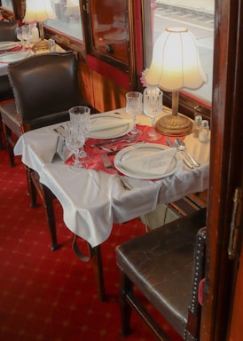 A cozy dining setup inside a vintage train car with a beautifully set table adorned with fine china, crystal glassware, and polished silverware. A warm, soft light radiates from a classic table lamp, creating an intimate and elegant atmosphere. The red carpeting adds a touch of luxury, accentuated by the dark leather chairs.