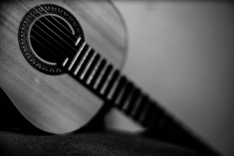A close-up shot of a classical guitar is captured in black and white, with sharp focus on the intricate soundhole design and blurred frets extending into the distance. The image emphasizes the textures of the wood and strings.