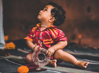 A young child wearing a colorful, traditional outfit is seated on a patterned floor. The child holds a silver object and looks upwards with curiosity. There is a bright orange marigold flower and red fabric near the child on the floor, adding to the cultural setting.