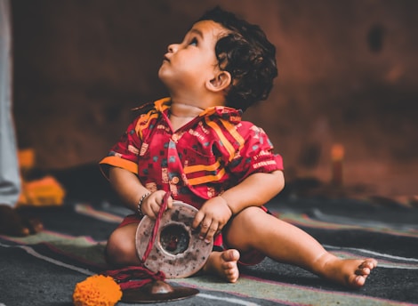 A young child wearing a colorful, traditional outfit is seated on a patterned floor. The child holds a silver object and looks upwards with curiosity. There is a bright orange marigold flower and red fabric near the child on the floor, adding to the cultural setting.