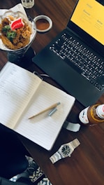 A wooden table holds a notebook with handwritten notes and a pencil, a laptop with a presentation slide open, a bottle of drink, a wristwatch, and a bowl of fried chicken with sauce. The setting suggests a combination of work and leisure.