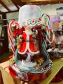 A colorful Christmas-themed decoration features a detailed figure of Santa Claus in a red suit holding a small lantern. Santa is surrounded by candy canes and miniature wrapped presents. The base includes intricate patterns with red and white swirls and holiday motifs.