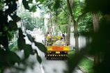 A work truck with a sign reading 'Work in Progress' is parked on a residential street surrounded by lush greenery. The street is lined with tall trees and there are buildings in the background. The scene is tranquil and partially obscured by leaves, suggesting a quiet, urban environment.