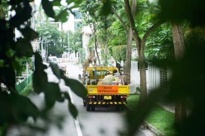 A work truck with a sign reading 'Work in Progress' is parked on a residential street surrounded by lush greenery. The street is lined with tall trees and there are buildings in the background. The scene is tranquil and partially obscured by leaves, suggesting a quiet, urban environment.