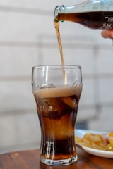 A glass is filled with a dark beverage being poured from a bottle. The drink is fizzy, with some ice cubes visible in the glass. In the background, there's a plate with snacks on a wooden surface.