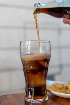 A glass is filled with a dark beverage being poured from a bottle. The drink is fizzy, with some ice cubes visible in the glass. In the background, there's a plate with snacks on a wooden surface.