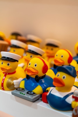 A collection of rubber duck toys, each styled in unique costumes. One duck is dressed as a DJ with headphones and a mixing console, while others are dressed as sailors and workers with hats.