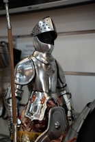 An armored figure in shiny metallic plate armor stands upright, displaying intricate details on the chestplate and including leather straps and decorative fabric elements at the waist. The helmet is perforated with holes and the visor is down, obscuring any face. A wooden staff with a metallic top is held by the figure. The background includes part of another armor set and a white wall with some indistinguishable text.
