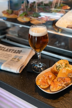 A glass of beer with a foamy head is placed on a dark counter next to a dish of assorted tapas, which includes slices of bread topped with different ingredients like smoked salmon and herbs. Nearby, a folded newspaper rests, lending a casual, leisurely atmosphere.