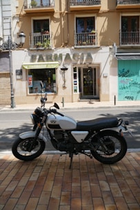 A vintage-style motorcycle is parked in front of a pharmacy. The building has a beige facade with a large sign reading 'FARMACIA' and two windows with decorative railings, each containing plants. Graffiti is visible on a closed storefront to the right. The ground is covered in light brown tiles.