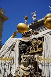 An elaborate and artistic sculpture featuring a combination of human busts and figures intricately carved in relief. The centerpiece is a bust of a bearded man, surrounded by detailed sculptures depicting various scenes. The structure is adorned with large golden spheres and draped with white fabric, set against the backdrop of classical architecture under a clear blue sky.