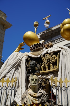 An elaborate and artistic sculpture featuring a combination of human busts and figures intricately carved in relief. The centerpiece is a bust of a bearded man, surrounded by detailed sculptures depicting various scenes. The structure is adorned with large golden spheres and draped with white fabric, set against the backdrop of classical architecture under a clear blue sky.