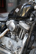 A close-up view of a Harley-Davidson motorcycle engine with visible details of the chrome components, including the air filter, exhaust pipes, and cylinder heads. The black gas tank prominently displays the Harley-Davidson logo, reflecting light and surrounding environment.