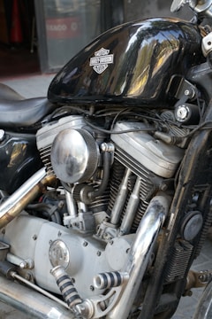 A close-up view of a Harley-Davidson motorcycle engine with visible details of the chrome components, including the air filter, exhaust pipes, and cylinder heads. The black gas tank prominently displays the Harley-Davidson logo, reflecting light and surrounding environment.