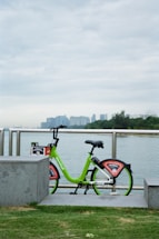 A green rental bicycle is parked beside a metal railing overlooking a body of water. In the background, there is a city skyline with tall buildings partially obscured by clouds. The scene includes concrete blocks and a grassy patch in the foreground.