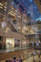 A bustling shopping mall is adorned with festive decorations, including illuminated snowflakes and cascading lights hanging from the tall ceiling. People are walking around on multiple floors, with various stores visible, including one with a visible Black Friday sale sign.