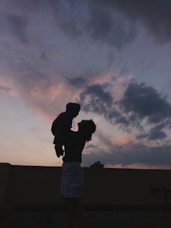 A silhouette of a person holding up a child against a dramatic sky filled with soft clouds that are tinged with shades of pink and purple.