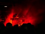 Dark stage with intense red lighting surrounding musicians playing electronic instruments, creating a moody and atmospheric setting. Silhouettes of performers are visible behind a veil of smoke or haze, heightening the dramatic effect.