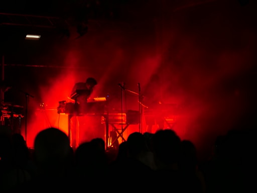 Dark stage with intense red lighting surrounding musicians playing electronic instruments, creating a moody and atmospheric setting. Silhouettes of performers are visible behind a veil of smoke or haze, heightening the dramatic effect.