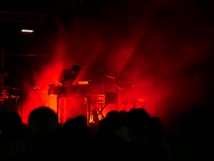 Dark stage with intense red lighting surrounding musicians playing electronic instruments, creating a moody and atmospheric setting. Silhouettes of performers are visible behind a veil of smoke or haze, heightening the dramatic effect.