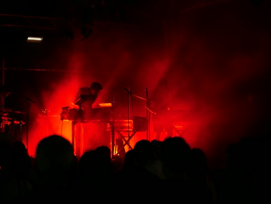 Dark stage with intense red lighting surrounding musicians playing electronic instruments, creating a moody and atmospheric setting. Silhouettes of performers are visible behind a veil of smoke or haze, heightening the dramatic effect.