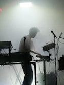 A silhouette of a person engaged in a live electronic music performance, surrounded by various sound equipment such as keyboards, synthesizers, and microphones. The stage is filled with fog, creating a moody and atmospheric ambiance. A strong light from above adds to the dramatic effect.