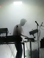 A silhouette of a person engaged in a live electronic music performance, surrounded by various sound equipment such as keyboards, synthesizers, and microphones. The stage is filled with fog, creating a moody and atmospheric ambiance. A strong light from above adds to the dramatic effect.